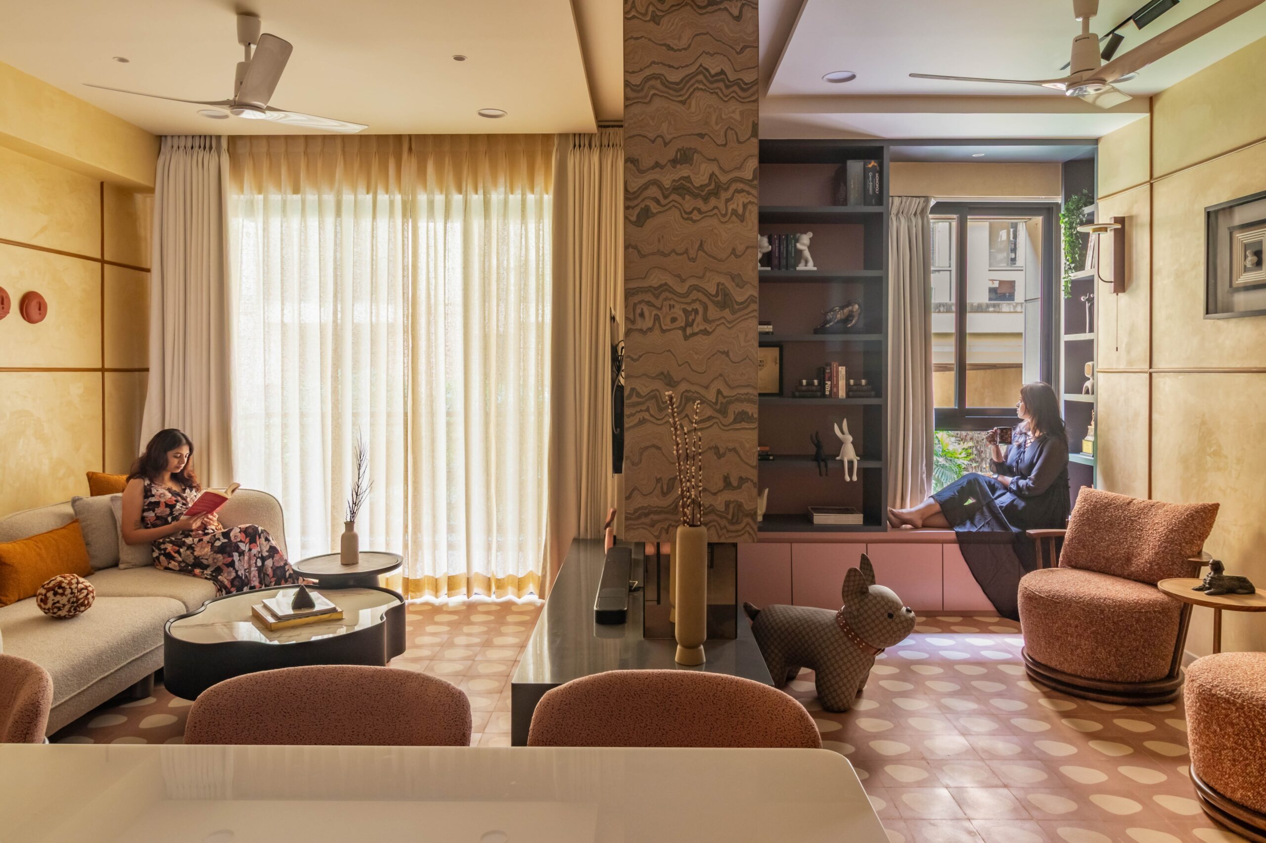 Warm contemporary Indian living room featuring a cozy seating area, large sheer curtains, custom window seat, textured wall finishes, designer furniture, and abundant natural light.