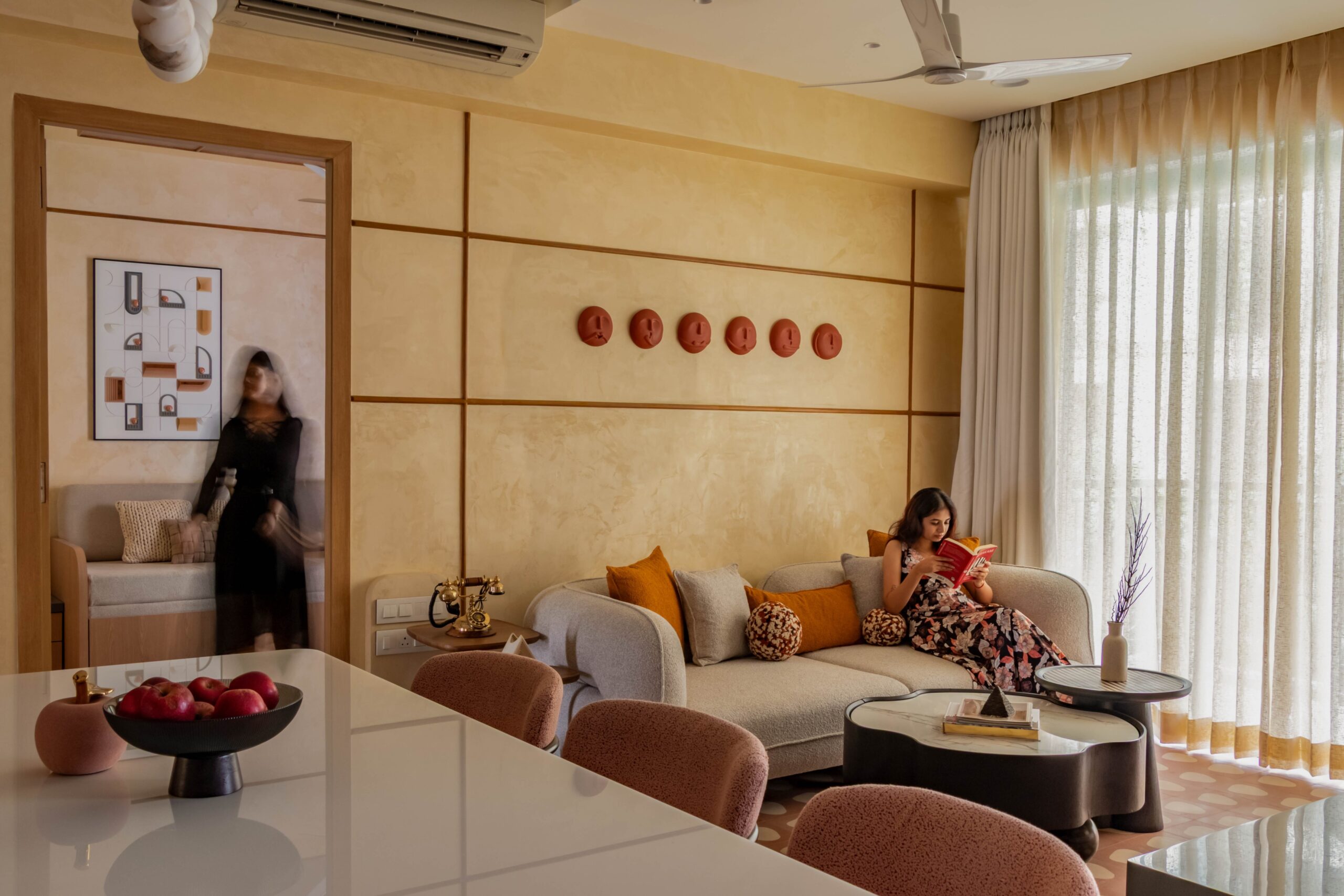 Cozy modern living room featuring a curved sofa, large floor-to-ceiling windows with sheer curtains, designer coffee table, warm textured walls, and a comfortable reading corner.