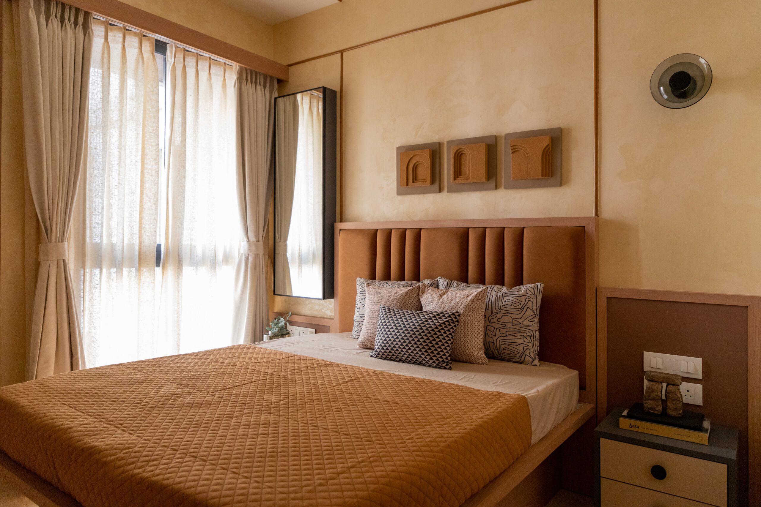 Warm contemporary Indian bedroom with a brown upholstered headboard, textured walls, large window with sheer curtains, wooden furniture, and minimalist décor.